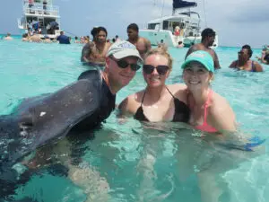 Family visiting Stingray City in Grand Cayman one of the Best Western Caribbean cruise excursions. With a stingray resting on man’s back in shallow water.