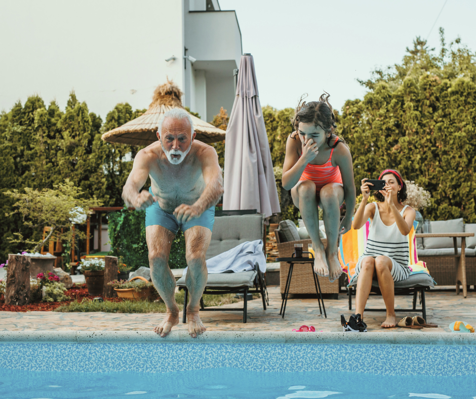 Grandpa and granddaughter jumping into a pool with a mom in the background capturing the moment with a picture.